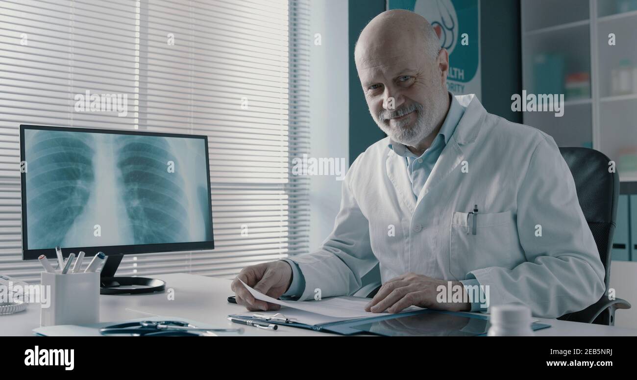 Medico professionista che lavora nel suo ufficio, sta controllando le cartelle cliniche e sorridendo Foto Stock