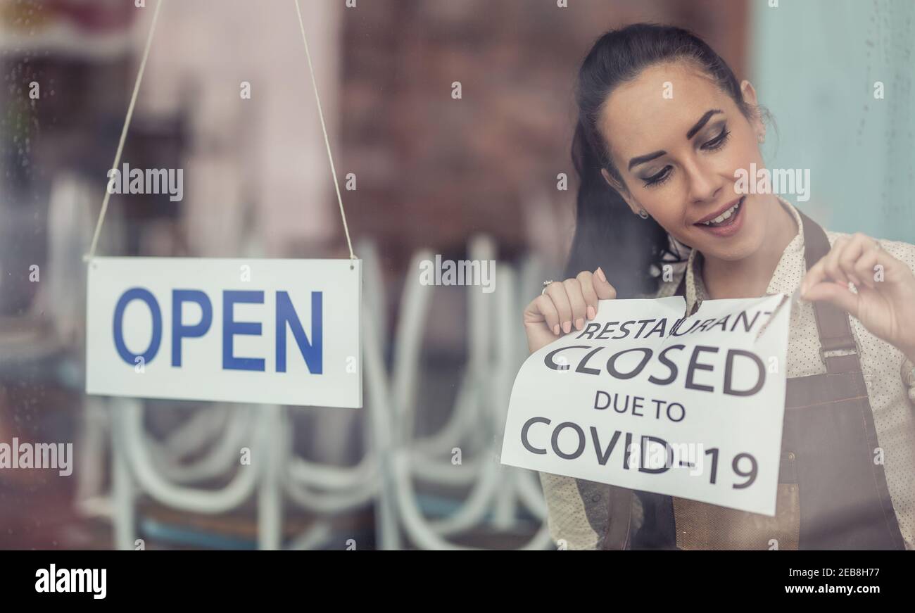 Come Covid-19 restrizioni facilità, il proprietario del ristorante sfila il segno di chiusura mettendo il segno aperto su una finestra. Foto Stock