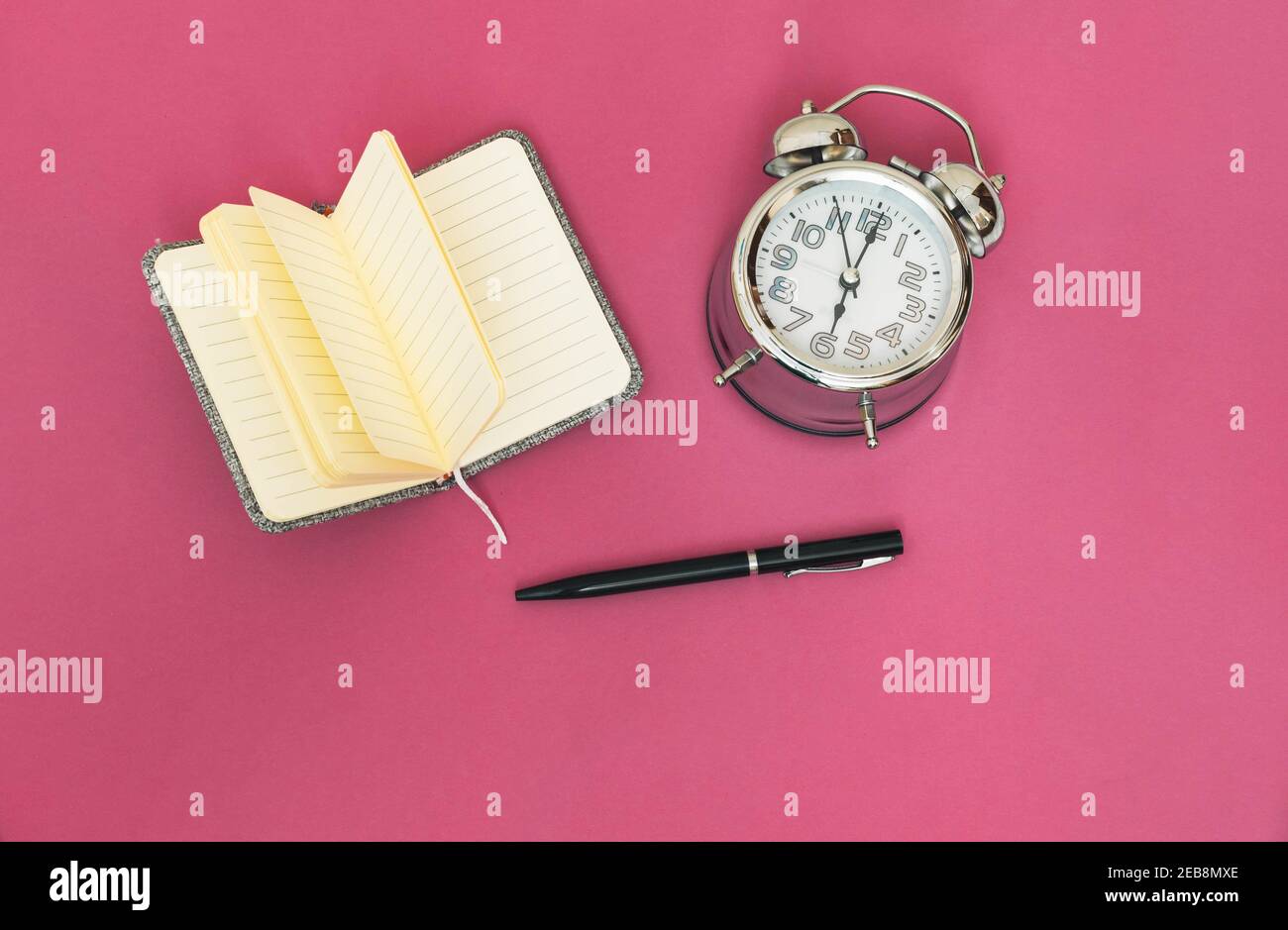 Vista dall'alto di un notebook aperto per le note di progetto, una penna nera e un orologio analogico per idee su sfondo rosa. Foto Stock