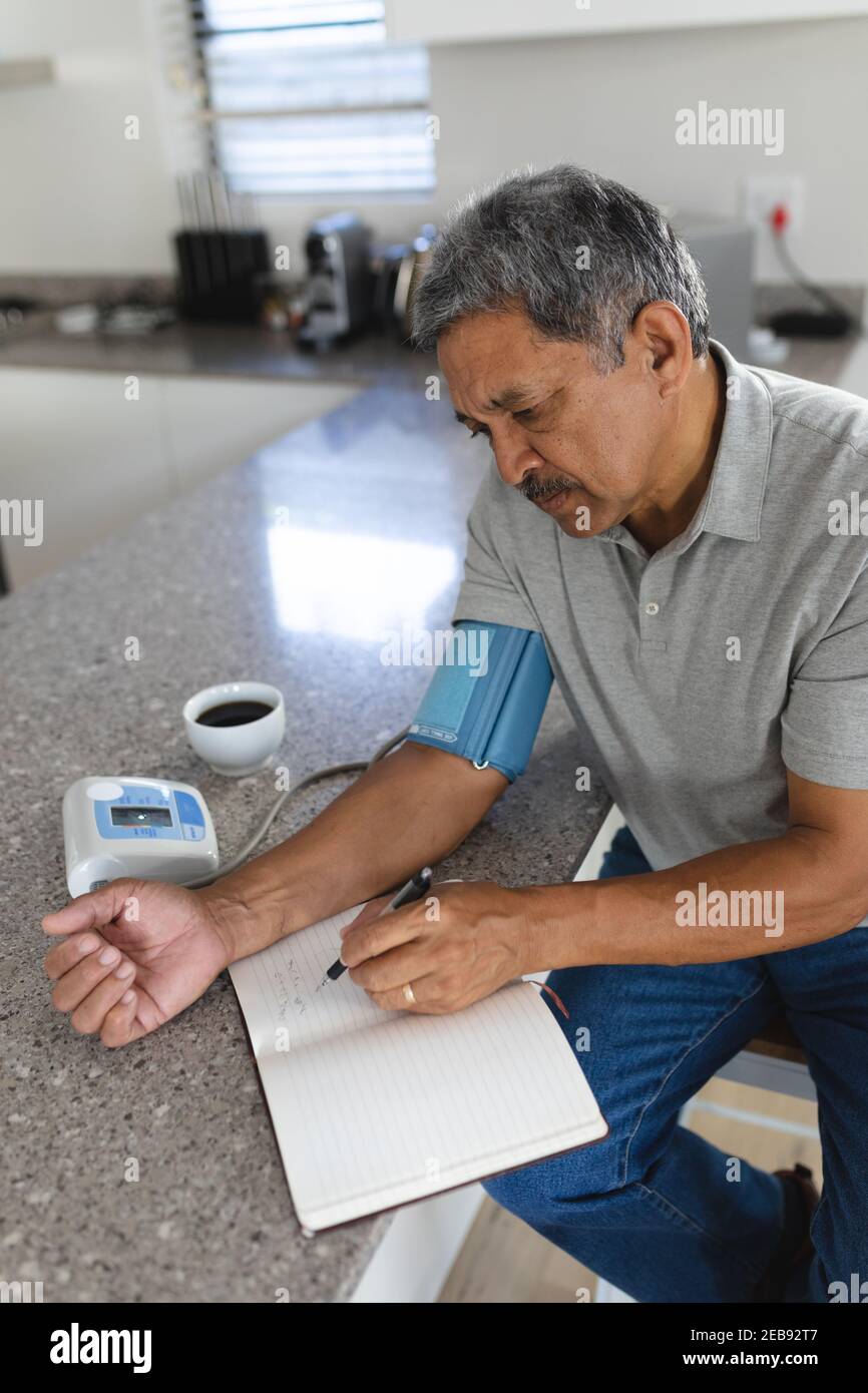 Uomo di razza misto senior a casa prendendo la sua pressione sanguigna e annotarlo Foto Stock