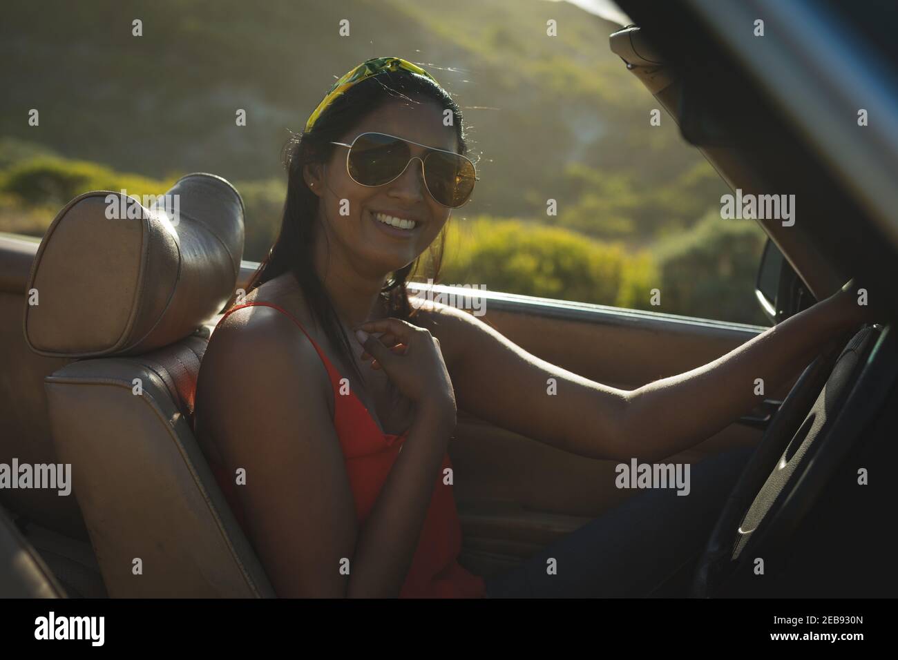 Donna mista che guida il giorno di sole in auto convertibile tenendo la ruota motrice e sorridendo Foto Stock