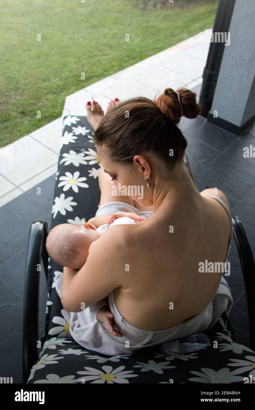 Vista posteriore della donna che tiene il bambino tra le braccia della madre e allatta al seno, mamma con un bambino seduto sulla sedia a sdraio del loggiato Foto Stock