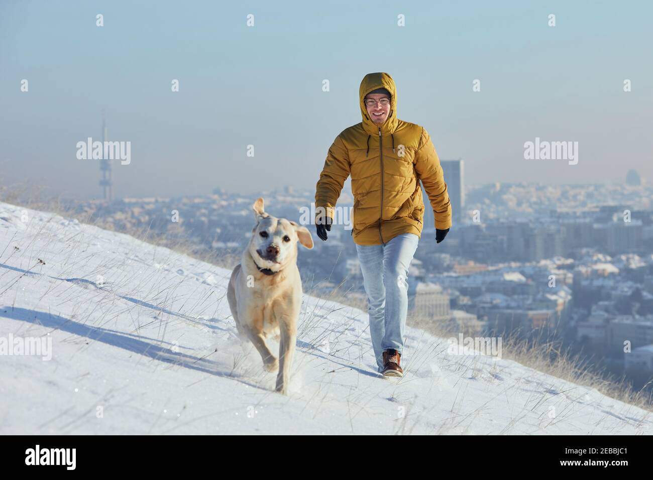 Felice giovane uomo con cane in inverno. Proprietario di animali domestici con il suo labrador Retriever che cammina nella neve contro la città. Praga, Repubblica Ceca Foto Stock