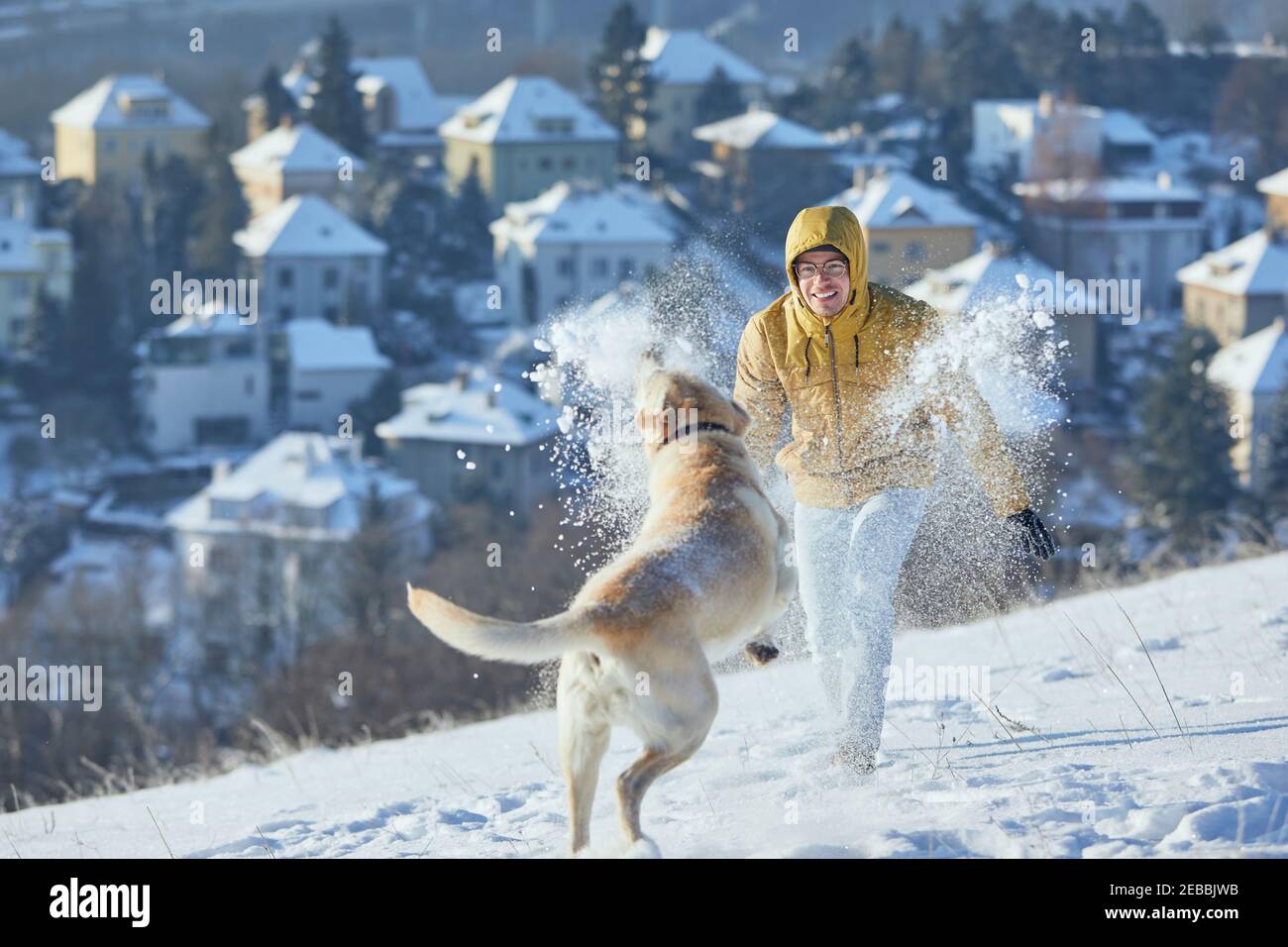 Giovane uomo con cane in inverno. Proprietario di animali domestici con il suo labrador Retriever che gioca nella neve contro la città. Praga, Repubblica Ceca Foto Stock