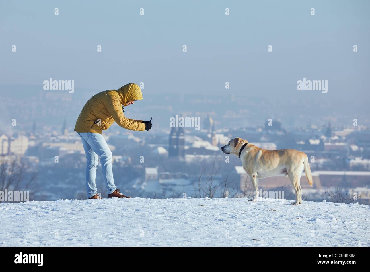 Il proprietario del PET scatta foto del suo labrador Retriever tramite smartphone durante la giornata gelosa. Giovane uomo con cane in inverno contro lo skyline urbano. Praga, ceco Foto Stock