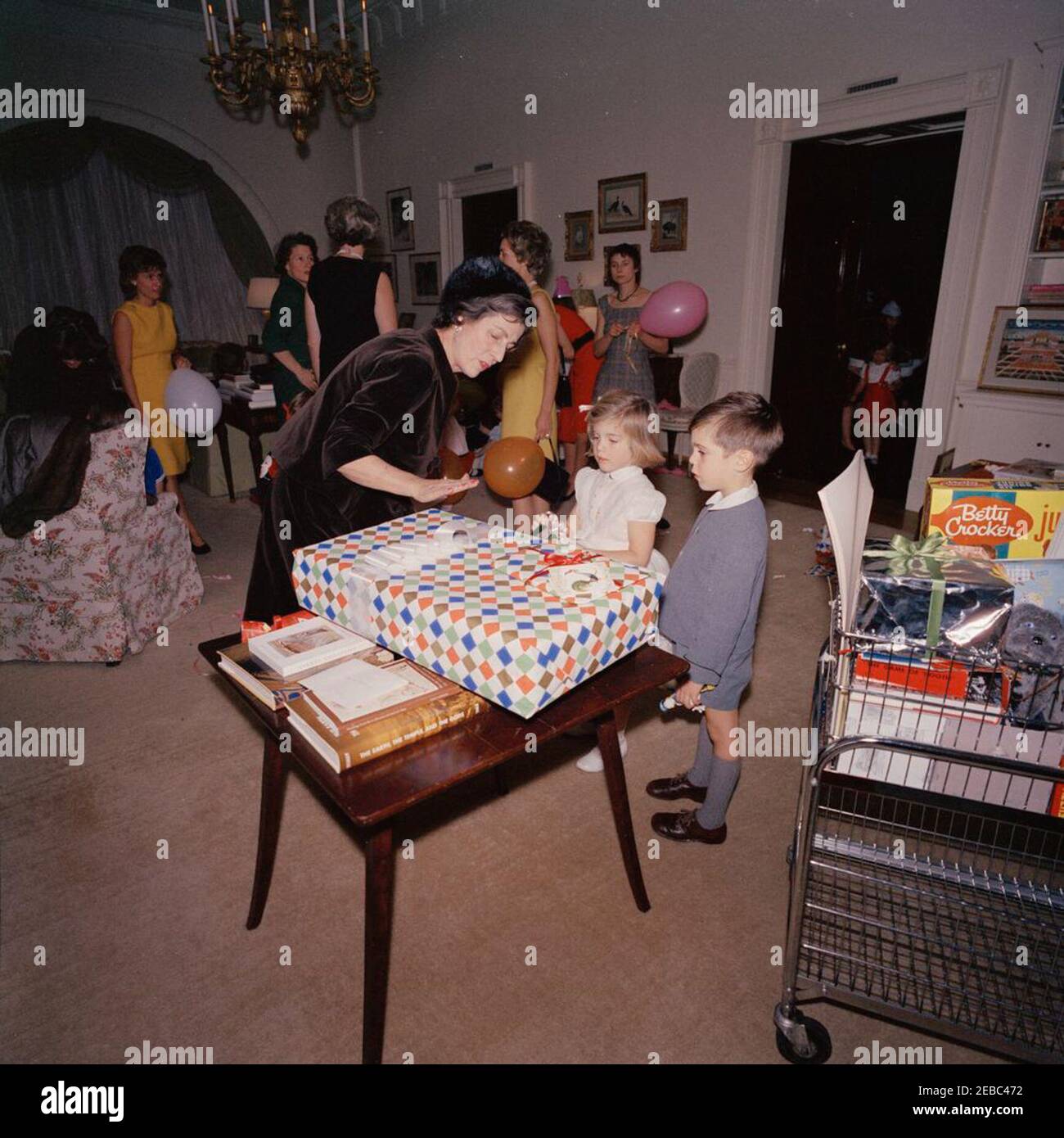 Festa di compleanno per Caroline Kennedy e John F. Kennedy, Jr. Caroline Kennedy visita con sua nonna, Janet Auchincloss, durante una festa di compleanno congiunta per lei e suo fratello, John F. Kennedy, Jr.; diversi regali siedono sul tavolo in primo piano. Anche nella foto: Eunice Kennedy Shriver; Maria Shriver; primo Segretario sociale Ladyu0027s, Letitia Baldrige. West sitting Hall, Casa Bianca, Washington, D.C. Foto Stock