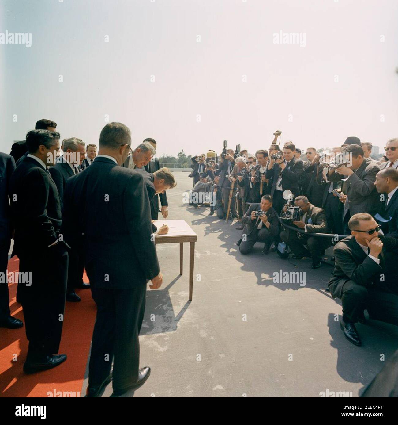 Viaggio negli Stati occidentali: Los Angeles, California, arrivo al Beverly Hilton Hotel per una cena di raccolta fondi, 15:40. Il presidente John F. Kennedy (appoggiandosi al tavolo al centro, tenendo una penna) firma un documento dopo il suo arrivo all'eliporto sul tetto dell'hotel Beverly Hilton a Los Angeles, California; il presidente Kennedy si è recato a Los Angeles per partecipare ad una cena di raccolta fondi del Partito democratico. Il Senatore Clair Engle (California) si trova sulla moquette rossa a sinistra; l'agente del Servizio segreto della Casa Bianca, Dick Johnsen, si inginocchia all'estrema destra. Il fotografo della Casa Bianca, Captain Cecil Stoughton (fotocamera), si inginocchia Foto Stock