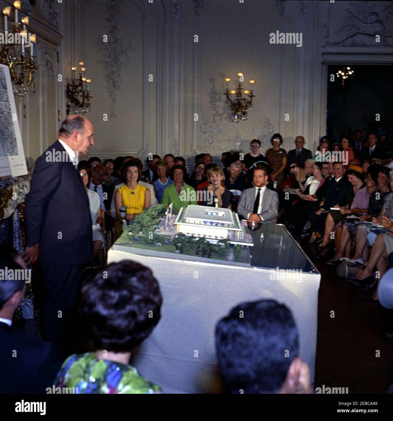 First Lady vede il modello del Centro Culturale Nazionale di Newport, Rhode Island. L'architetto, Edward Durell Stone, parla alla presentazione di un modello architettonico del Centro Culturale Nazionale in una cerimonia presso il palazzo Elms a Newport, Rhode Island; First Lady Jacqueline Kennedy (vestita gialla) si trova in prima fila dietro il modello. Anche nella foto: presidente del Washington Area Campaign Committee per il Centro Culturale Nazionale, Janet Auchincloss (parzialmente oscurato); direttore musicale della Boston Symphony Orchestra, Erich Leinsdorf; membro del Comitato consultivo per le Arti f Foto Stock
