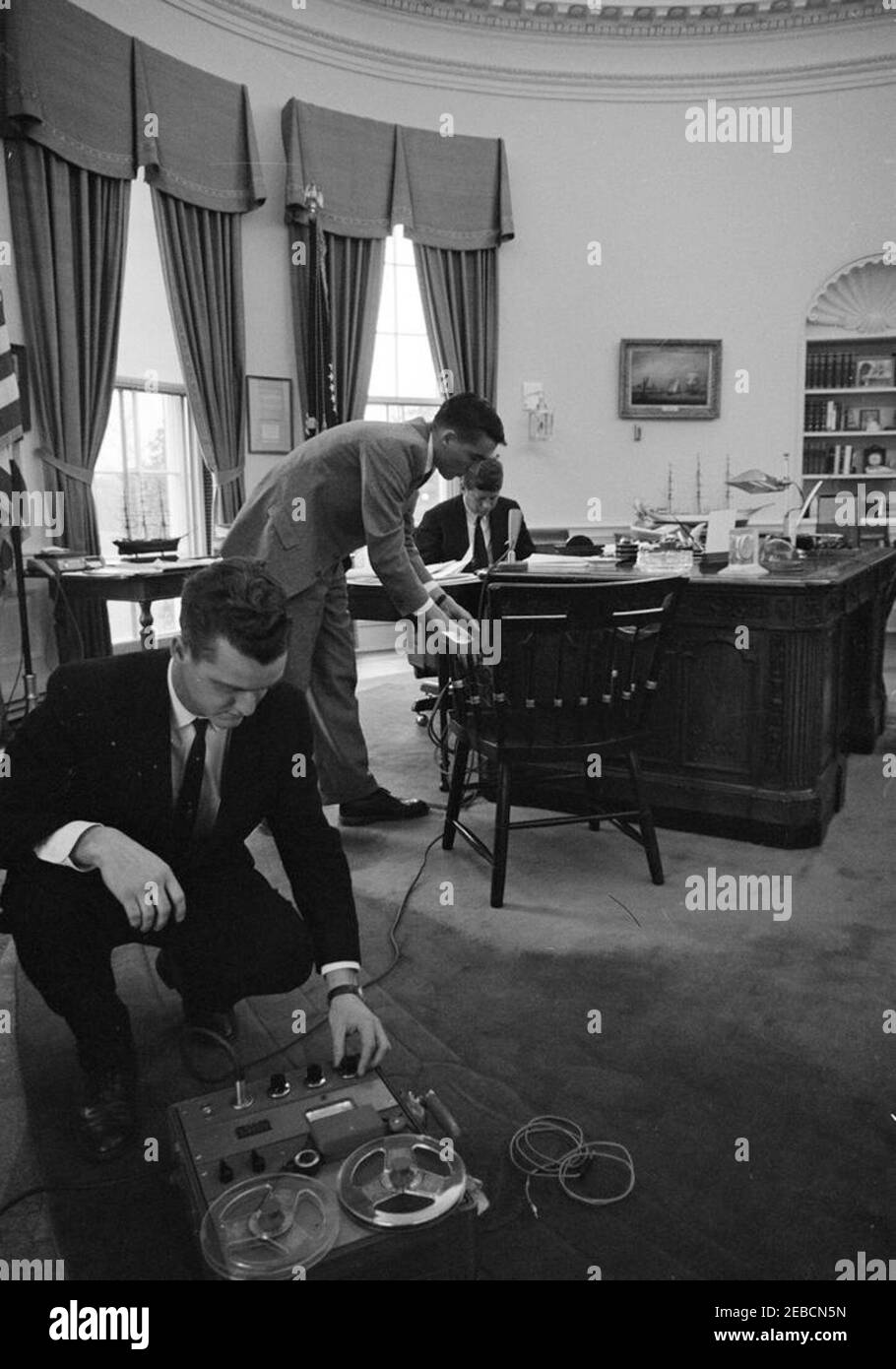 Osservazioni registrate per l'appello annuale della Croce Rossa americana. Il presidente John F. Kennedy (seduto alla scrivania) si prepara a registrare una dichiarazione per la trasmissione televisiva e radiofonica sull'appello annuale per la Croce Rossa americana. White House Army Signal Agency (WHASA) membri dello staff Oscar N. Smith (primo da sinistra) e John J. Cochran (in salita) hanno allestito attrezzature in primo piano. Ufficio ovale, Casa Bianca, Washington, D.C. Foto Stock