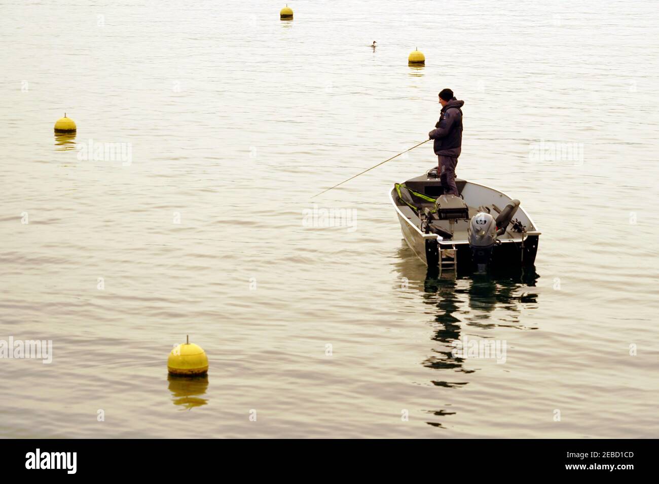 Pescatore hobby in piedi su una barca a motore che tiene una canna da pesca sul lago di Zurigo in inverno. Ci sono boe gialle intorno e molto spazio di copia. Foto Stock