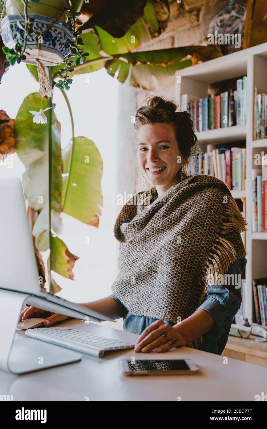 Donna lavora su un computer portatile dall'ufficio domestico circondato da libri e piante Foto Stock