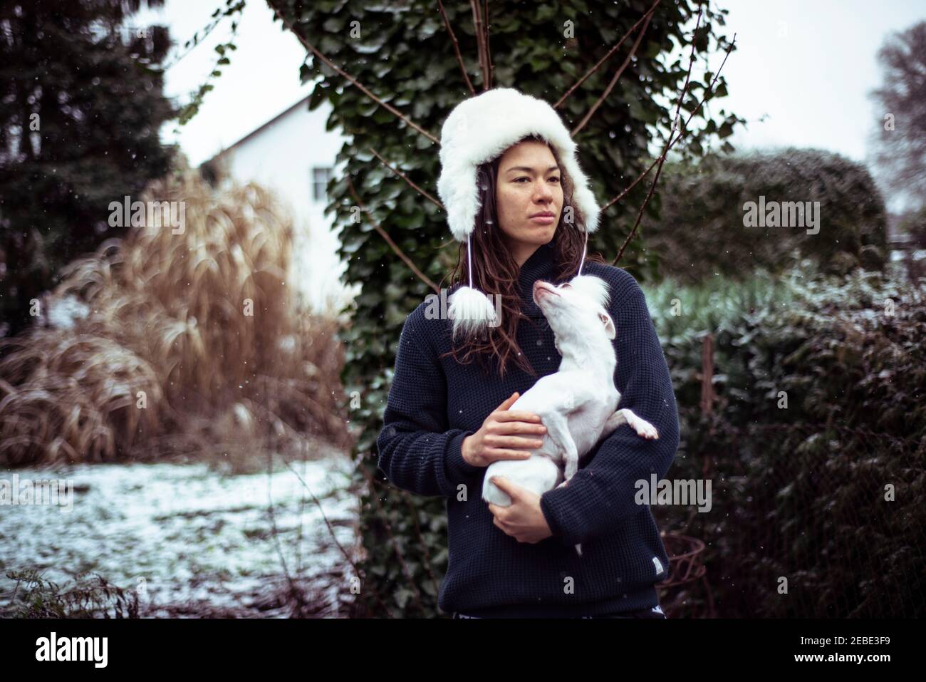 Persona in bianco morbido cappello si alza in neve con piccolo cane bianco in cortile Foto Stock