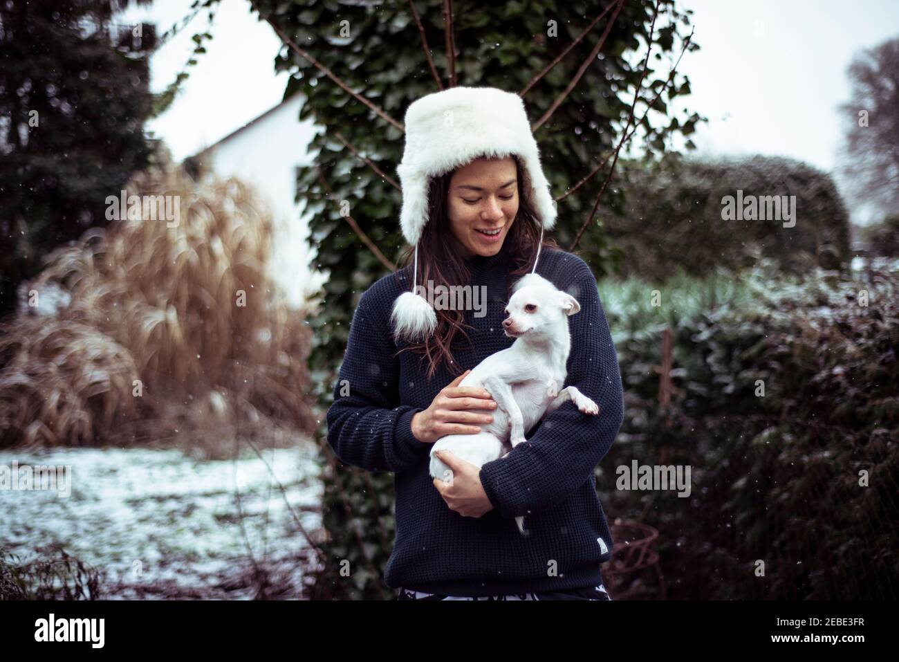 La donna di razza mista sorride al cane bianco in armi nella neve e cappello morbido Foto Stock