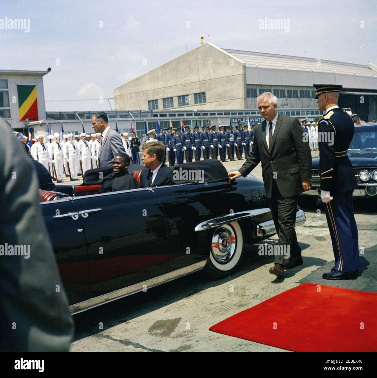 Cerimonia di arrivo per Fulbert Youlou, Presidente della Repubblica del Congo (Brazzaville), ore 12. Cerimonia di arrivo per il Presidente della Repubblica del Congo (Brazzaville) Fulbert Youlou. Il presidente Youlou e il presidente John F. Kennedy cavalcano sul sedile posteriore di un'automobile de Lincoln (convertibile). Roy Kellerman, agente di servizio segreto della Casa Bianca, si trova a sinistra dietro l'auto. Guardie d'onore per la Marina militare degli Stati Uniti e l'Aeronautica militare sono sullo sfondo. Terminal dei servizi di trasporto aereo militare (MATS), Washington National Airport, Washington, D.C. Foto Stock