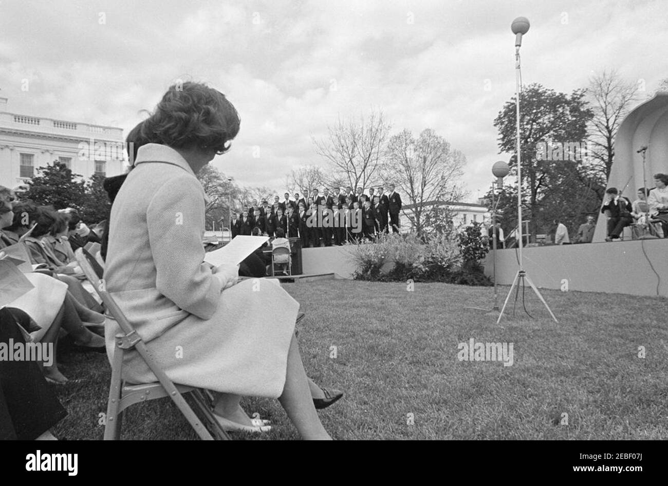 First Lady Jacqueline Kennedyu0027s (JBK) programma musicale per la gioventù, 14:35. First Lady Jacqueline Kennedy (a sinistra, di nuovo alla telecamera) guarda dalla prima fila del pubblico come direttore del coro, Gwen Dean (seduto a sinistra al piano), conduce una performance del coro Breckenridge Boys durante la terza nella serie di programmi musicali per giovani della signora Kennedyu0027s. I membri della Greater Boston Youth Symphony Orchestra (GBYSO) siedono sul palco all'estrema destra. South Lawn, Casa Bianca, Washington, D.C. Foto Stock