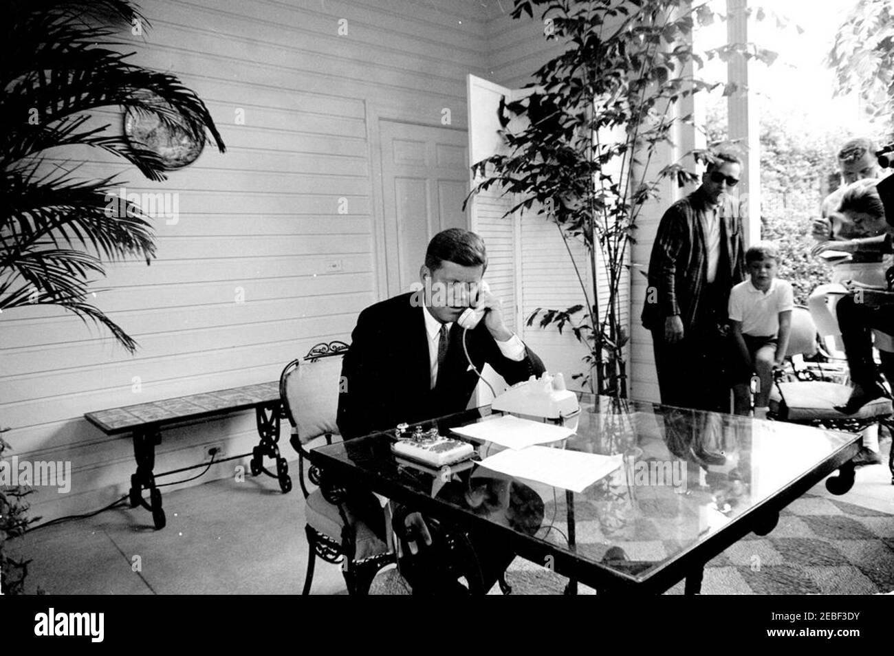 Cerimonia di apertura della fiera di Seattle Worldu0027s da Palm Beach. Il presidente John F. Kennedy rilascia le sue osservazioni per telefono prima di premere un tasto del telegrafo d'oro (sul tavolo del patio) per aprire la fiera di Seattle Worldu0027s del 1962 (conosciuta anche come esposizione del secolo 21) via segnale satellitare dalla residenza di C. Michael Paul a Palm Beach, Florida. White House Secret Service Agent, Clint Hill (indossare occhiali da sole), si trova sullo sfondo a destra; tutti gli altri sono non identificati. Foto Stock