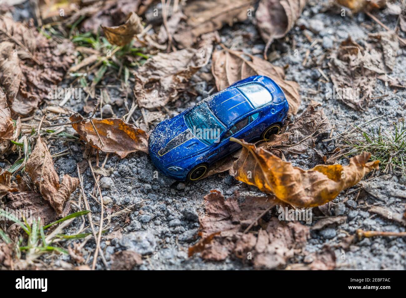 Una piccola auto giocattolo blu che si stesa al suolo coperta con sporcizia e sabbia circondati da foglie cadute a sinistra e. dimenticato lungo il sentiero in un parco al sole Foto Stock