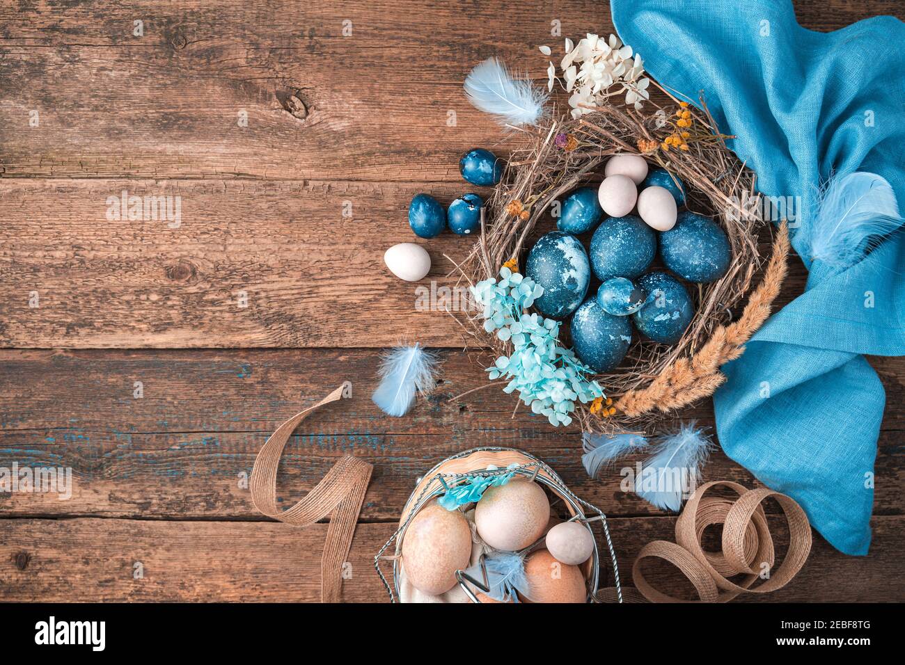 Uova di Pasqua blu-marmo in un nido di vimini con piume e fiori accanto a un tovagliolo blu e un cestino di uova pastello. Foto Stock