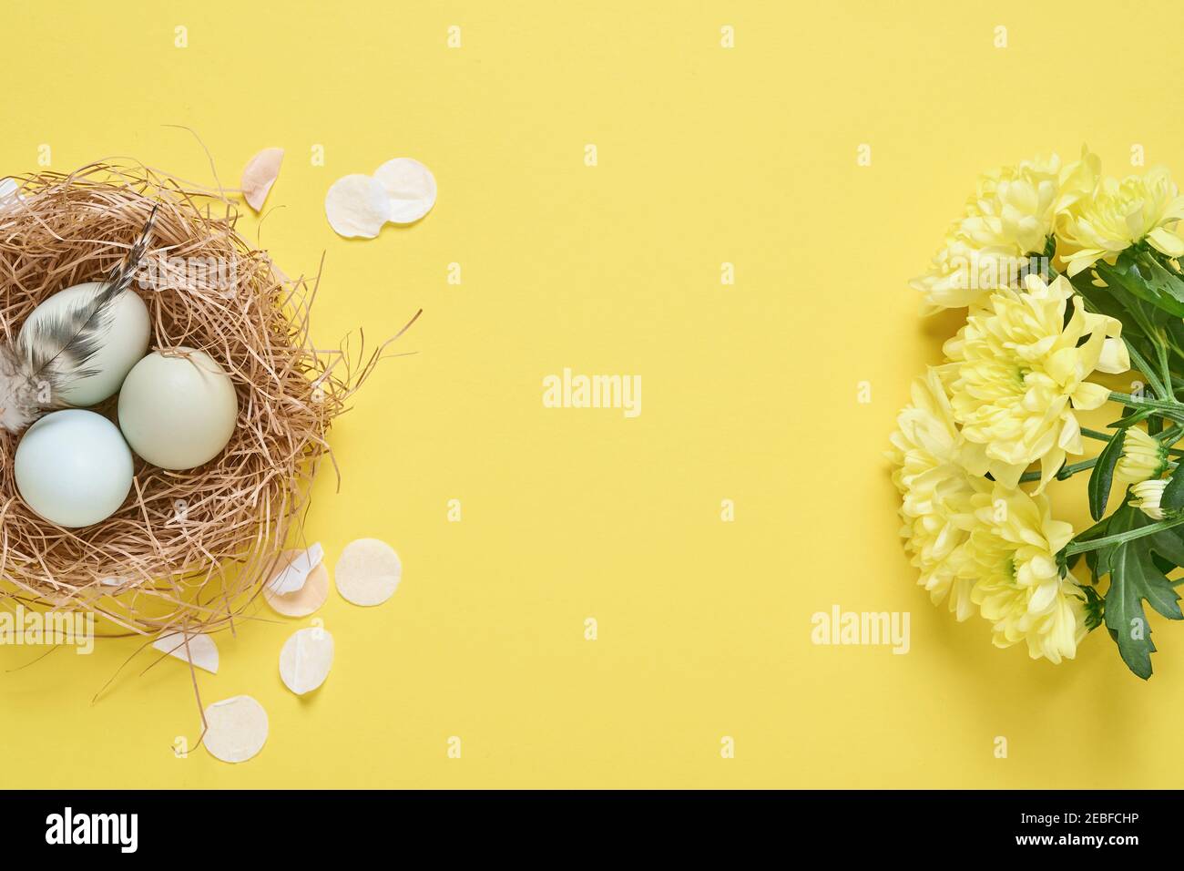 Uova blu chiaro di Pasqua in bianco vintage contenitore metallico con piume, nastro, fiori di crisantemi gialli e carta bianca per testo su sfondo giallo Foto Stock