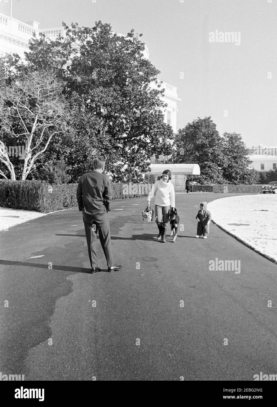 First Lady Jacqueline Kennedy (JBK) u0026 John F. Kennedy, Jr. (JFK, Jr.), sledding su South Lawn. First Lady Jacqueline Kennedy visita il presidente John F. Kennedy dopo aver dormito con John F. Kennedy, Jr., sul prato meridionale; il cane della famiglia Kennedy, Clipper, cammina accanto alla signora Kennedy. South Lawn vialetto, Casa Bianca, Washington, D.C. Foto Stock