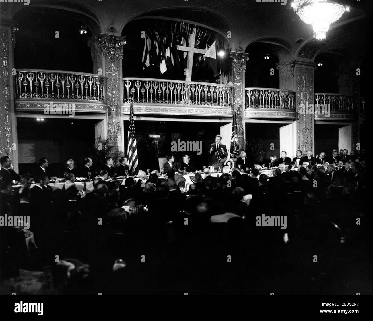 Commento alla colazione nazionale di preghiera 1961, 9:37. La colazione di preghiera sponsorizzata dal Consiglio Internazionale per la leadership cristiana nella grande sala da ballo, Mayflower Hotel, Washington, D.C. il reverendo Billy Graham si rivolge al pubblico dal leggio sotto una grande croce. (L-R) al tavolo: Tre uomini non identificati; ambasciatore degli Stati Uniti presso le Nazioni Unite Adlai E. Stevenson; Segretario della Difesa Robert S. McNamara; uomo non identificato; Presidente John F. Kennedy; co-presidente del senatore di prima colazione Frank Carlson del Kansas; reverendo Graham; Vice Presidente Lyndon B. Johnson; dieci non identificati Foto Stock