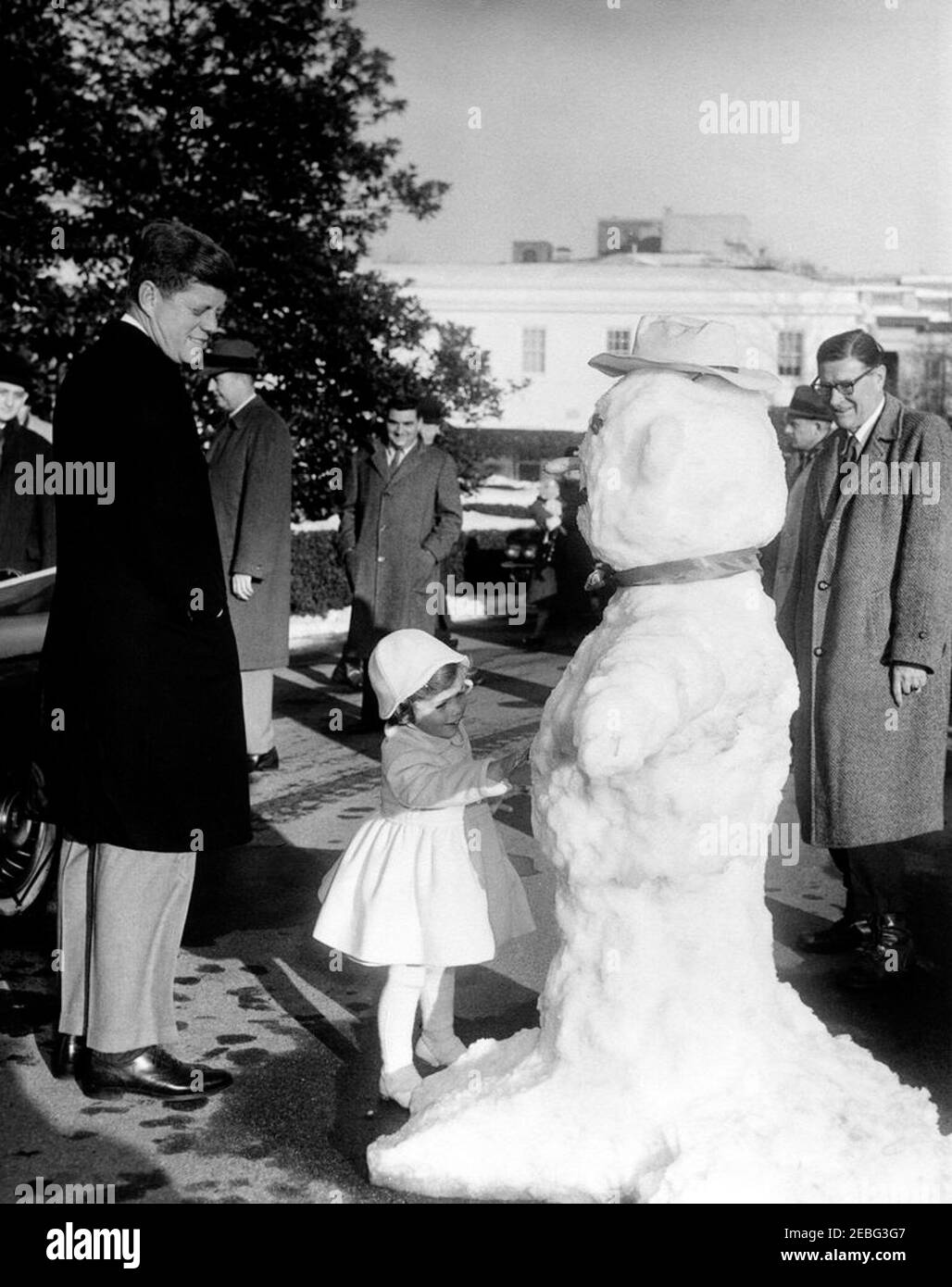 Caroline Kennedy (CBK) con un pupazzo di neve, 16:15. Il presidente John F. Kennedy guarda sua figlia Caroline mentre tocca un pupazzo di neve fatto per lei sul viale della Casa Bianca, Washington, D.C. il capo Gardener della Casa Bianca Robert M. Redmond si erge a destra del pupazzo di neve. White House Press Secretary Pierre Salinger orologi da dietro Caroline.rn Foto Stock