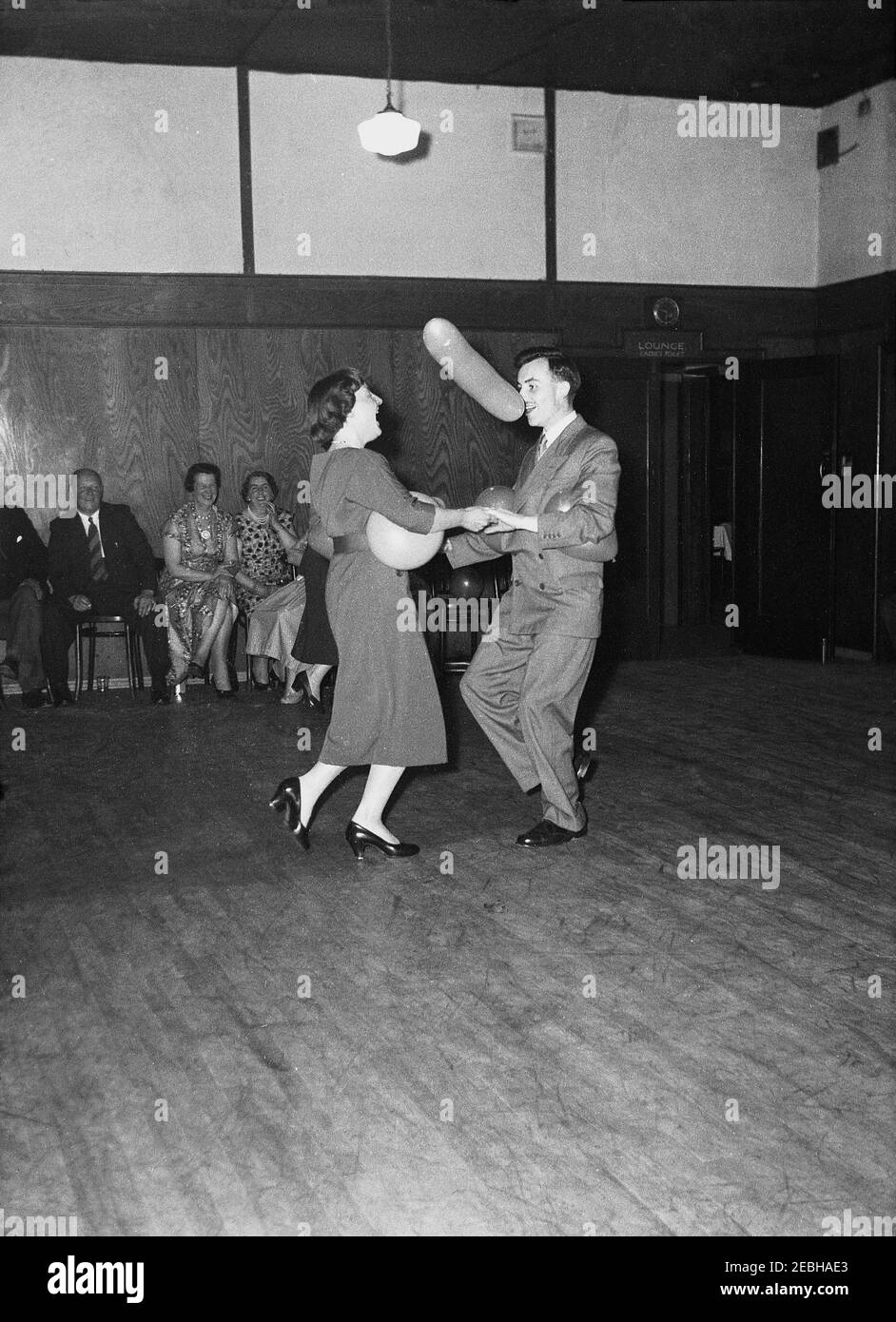 Anni '50, storico, risate tutto il giro dagli ospiti seduti come una coppia fa il ballon 'su una pista da ballo di legno ad una festa di compleanno in una stanza in un hotel, Inghilterra, Regno Unito. Qui il ballone è stato strofinato in modo che si tiene sul naso dell'uomo che balla, creando molto divertimento per la sua partner femminile sulla pista da ballo. Foto Stock