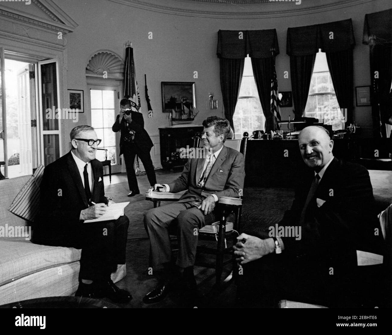 Incontro con Arthur A. Calwell, leader del Partito laburista d'Australia, ORE 12:05. Il presidente John F. Kennedy (in sedia a dondolo, tenendo una penna in una mano e un libro nell'altra) incontra il leader del Partito laburista australiano, Arthur A. Calwell (a sinistra, scrivendo in un libro); l'ambasciatore dell'Australia, Sir Howard Beale, siede a destra. Un fotografo non identificato (volto oscurato dalla fotocamera) scatta foto sullo sfondo a sinistra. Ufficio ovale, Casa Bianca, Washington, D.C. Foto Stock