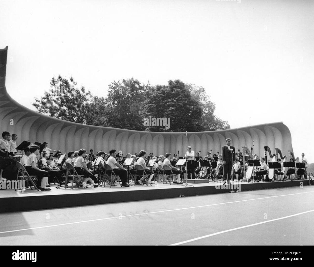 First Lady Jacqueline Kennedyu0027s (JBK) programma musicale per la gioventù, 10:35. Il presidente John F. Kennedy (a destra, ai microfoni) commenta la quarta serie di programmi musicali per giovani della First Lady Jacqueline Kennedyu0027s. I membri della National High School Symphony Orchestra del National Music Camp di Interlochen, Michigan, siedono sulla tribuna; direttore d'orchestra e presidente del National Music Camp, Dr. Joseph E. Maddy (armi incrociate), si trova a sinistra del presidente Kennedy. South Lawn, White House, Washington, D.C. [Ink bleed-through nella parte inferiore dell'immagine è Foto Stock