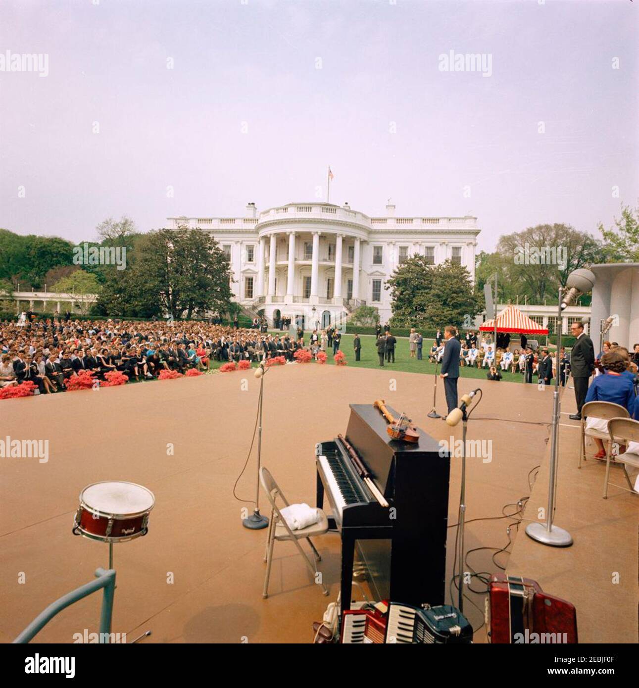 First Lady Jacqueline Kennedyu0027s (JBK) programma musicale per giovani, 14:10. Il presidente John F. Kennedy (in piedi ai microfoni) fa commenti durante una performance della Central Kentucky Youth Symphony Orchestra sul South Lawn della Casa Bianca, Washington, D.C.; il direttore Joseph Pival si trova sul palco all'estrema destra. I membri del Berea (Kentucky) College Country Dancer siedono sotto la tenda a strisce sullo sfondo a destra. I musicisti e i ballerini si sono esibiti nell'ambito della sesta puntata di First Lady Jacqueline Kennedyu2019s Musical Programs for Youth by Youth. Foto Stock