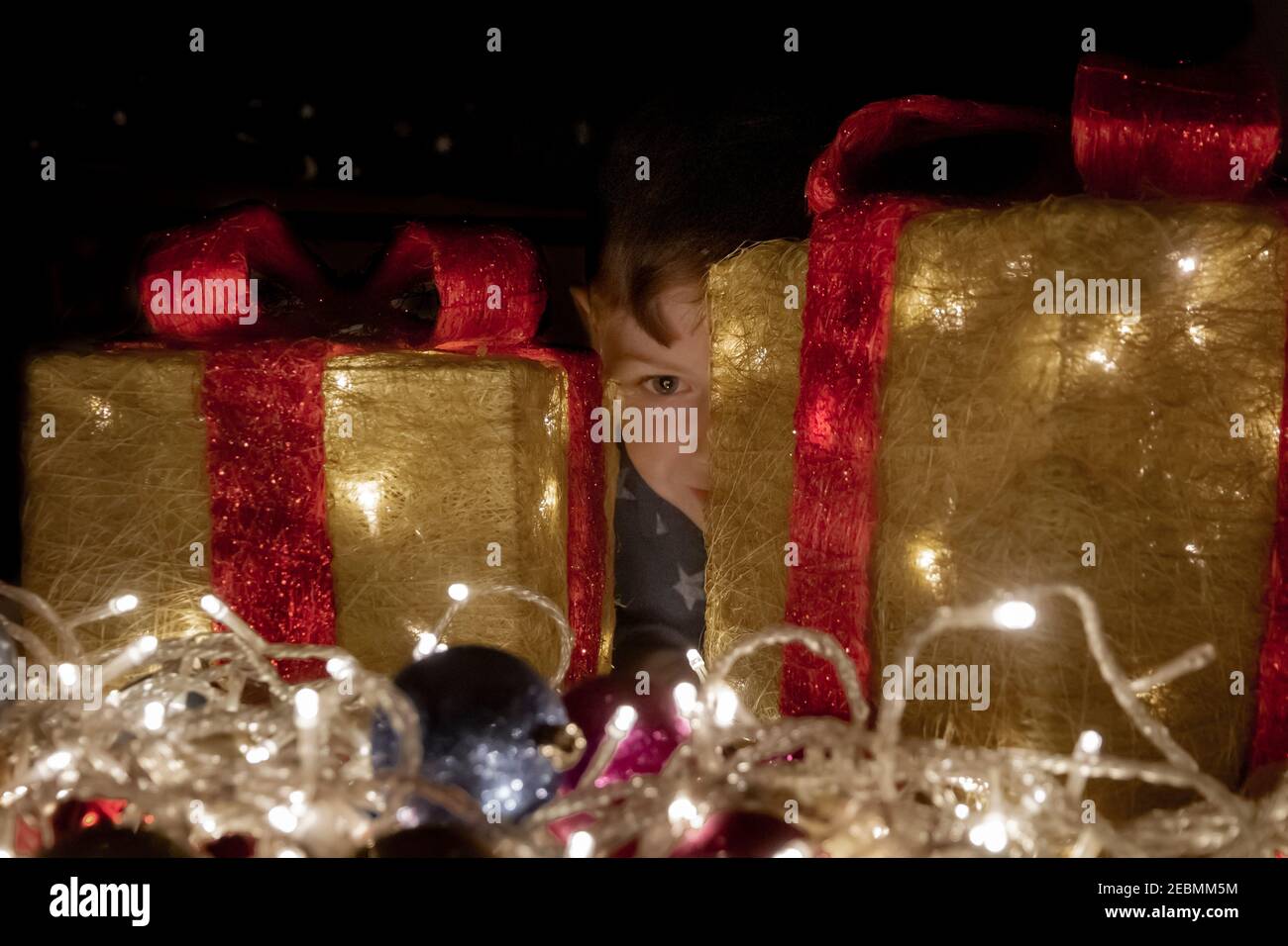 Ragazzo che si nasconde dietro qualche decorazione di Natale sotto forma di alcuni si presentano in una stanza buia Foto Stock