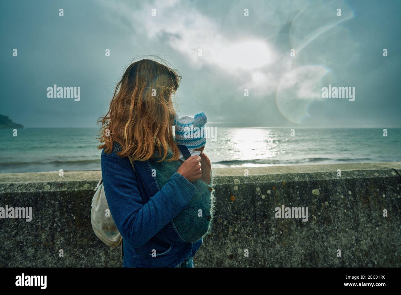 Una giovane madre con il suo bambino in un'imbragatura è in piedi sul mare e si affaccia su un muro Foto Stock