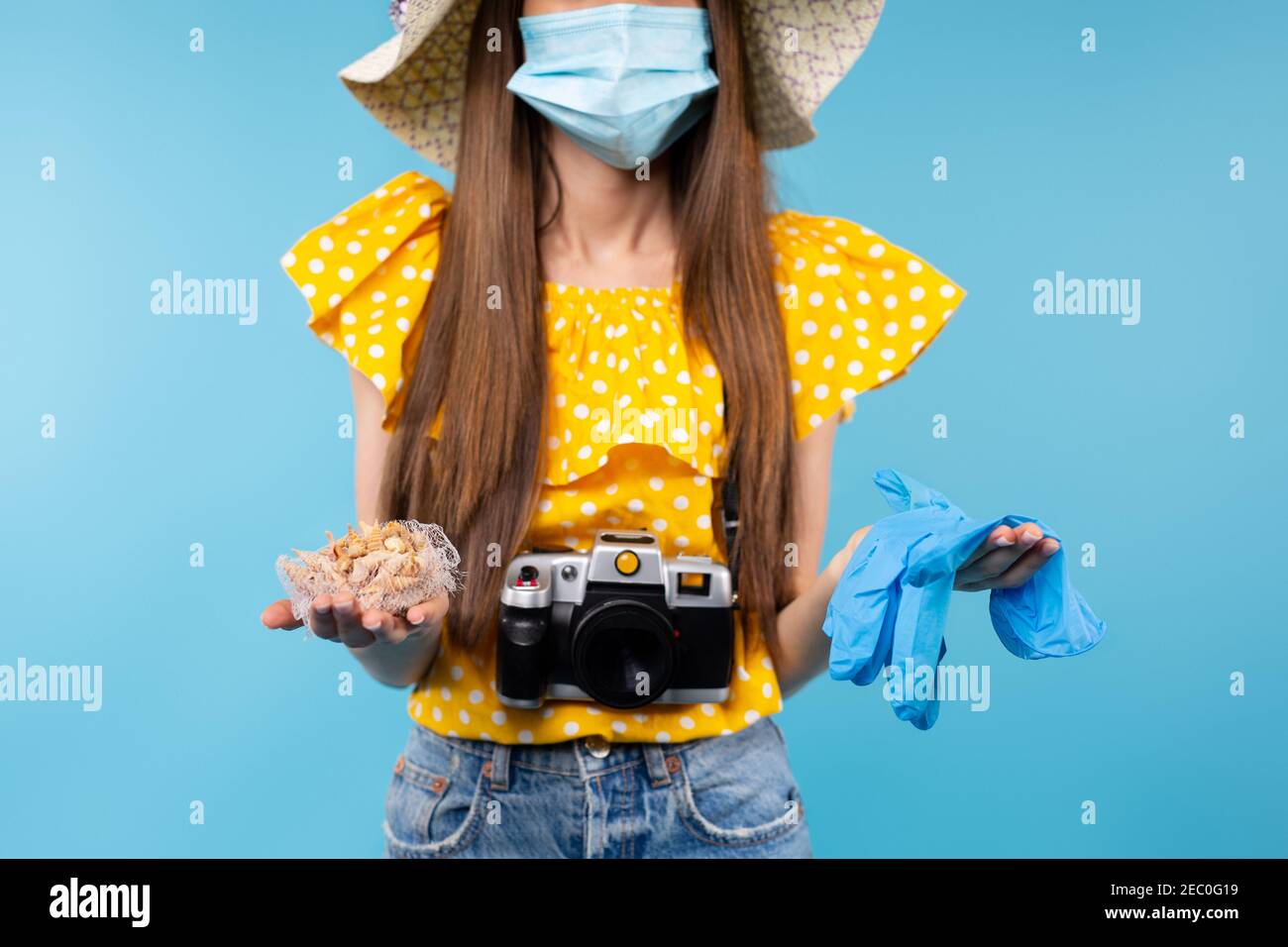 Foto ritagliata di donna brunetta con fotocamera che indossa maschera medica protettiva e cappello estivo, tenendo guanti medici blu e conchiglie marine. Pandemia Foto Stock