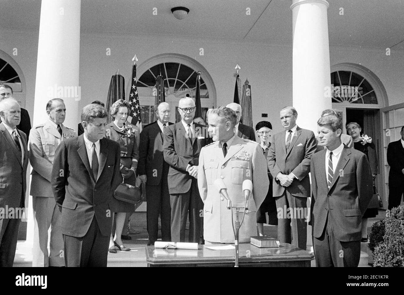 Cerimonia di giuramento, Gen. Maxwell Taylor, Presidente, JCS, 12:00. Il generale Maxwell D. Taylor (al centro a destra, ai microfoni), durante la sua cerimonia di giuramento come presidente dei capi di Stato e di Stato (JCS); il presidente John F. Kennedy (a sinistra) e il procuratore generale Robert F. Kennedy, si trovano su entrambi i lati del generale Taylor. Guardando dai gradini della West Wing Colonnade, a partire dalle spalle (da sinistra a destra): Naval Aide al presidente, Capitano Tazewell T. Shepard, Jr. (Parzialmente nascosto); Generale Omar Bradley; Presidente uscente della JCS, Generale Lyman L. Lemnitzer; Lydia Foto Stock