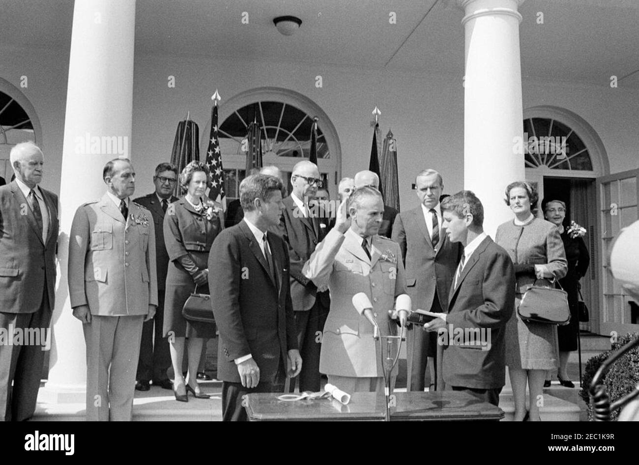 Cerimonia di giuramento, Gen. Maxwell Taylor, Presidente, JCS, 12:00. Il presidente John F. Kennedy (centro a sinistra) e altri osservano come il generale Maxwell D. Taylor (con la mano destra sollevata e la mano sinistra sulla Bibbia) è giurato come presidente dei capi di Stato Uniti (JCS); il procuratore generale, Robert F. Kennedy (centro a destra, tenendo la Bibbia), amministra il giuramento di carica. Guardando dai gradini della West Wing Colonnade (da sinistra a destra): Generale Omar Bradley; presidente uscente della JCS, generale Lyman L. Lemnitzer; capo di stato maggiore dell'aviazione militare degli Stati Uniti, generale Curtis E. LeMay; Lydia Taylor; se Foto Stock