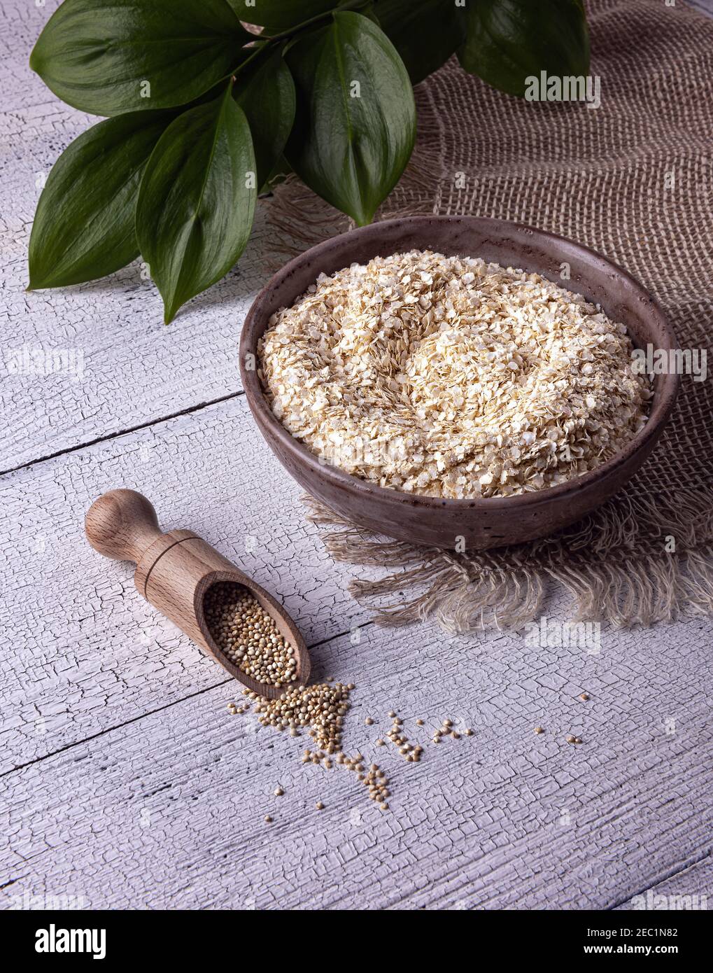 Scaglie di quinoa bianche grezze secche in ciotola di argilla fatta a mano su tovaglia di lino e paletta con semi di quinoa interi su tavolo di legno bianco. Foglie verdi A. Foto Stock