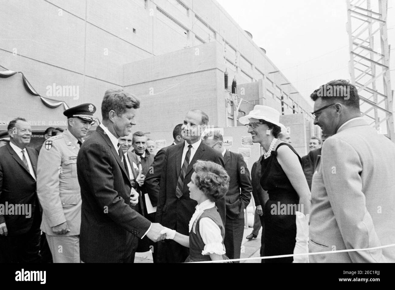 Viaggio negli stati occidentali: Pierre, South Dakota (Oahe Dam), 10:25. Il presidente John F. Kennedy scuote le mani con Jamie Damon, di Pierre, South Dakota, durante la cerimonia di dedicazione della diga e del serbatoio di Oahe, sulle rive del fiume Missouri, vicino a Pierre, South Dakota. Stati Uniti Senatorial candidato dal Dakota del Sud, George McGovern (centro), e Jamieu0027s madre, Dottie Damon (destra), sguardo sopra. Anche nella foto: Capo degli ingegneri militari, tenente generale Walter K. Wilson, Jr.; decano del corpo stampa della Casa Bianca e reporter della United Press International (UPI), Merriman u0022Smittyu0022 Smith. Foto Stock