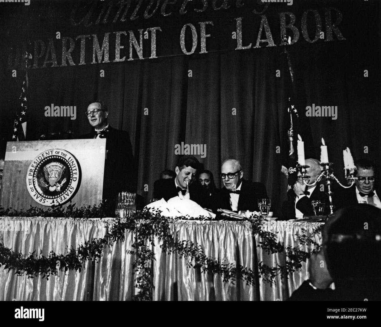 Discorso alla cerimonia del 50° anniversario del Dipartimento del lavoro, ore 10:26. Il presidente John F. Kennedy (fumando un sigaro) partecipa a una cena che celebra il 50° anniversario del Dipartimento del lavoro; il segretario del lavoro, W. Willard Wirtz, fa le sue osservazioni dal leggio. Seduto (L-R): Presidente Kennedy; Presidente della Federazione americana del lavoro e del Congresso delle organizzazioni industriali (AFL-CIO), George Meany; Postmaster Generale, J. Edward Day (dietro candelabra); ex Segretario del lavoro, James P. Mitchell. Sheraton Park Hotel, Washington, D.C. Foto Stock