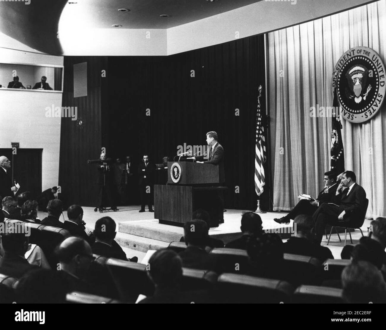 Conferenza stampa presso il Dipartimento di Auditorium di Stato, alle 18:00. Il presidente John F. Kennedy parla dal leggio durante una conferenza stampa. Il segretario alla stampa, Pierre Salinger, e il segretario aggiunto alla stampa, Malcolm Kilduff, siedono sul palco a destra. Auditorium del Dipartimento di Stato, Washington, D.C. Foto Stock