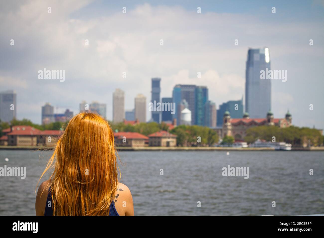 Una giovane donna con lunghi capelli rossi sta guardando il Skyline di Manhattan da Liberty Island a New York Foto Stock