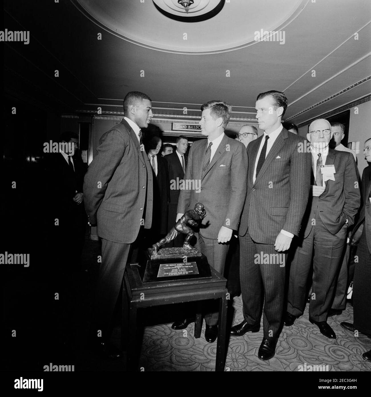 Il Presidente Kennedy saluta il vincitore del Trofeo Heisman 1961 Ernie Davis ad un ricevimento sponsorizzato dalla National Association of Manufacturers, Waldorf-Astoria Hotel, New York City, alle 12:07. Il presidente John F. Kennedy parla con il vincitore del Trofeo Heisman Ernie Davis (a sinistra) ad un ricevimento sponsorizzato dalla National Association of Manufacturers. White House Secret Service Agent Art Godfrey si trova dietro (a sinistra del presidente Kennedy); il presidente della National Association of Manufacturers John W. McGovern si trova all'estrema destra. Waldorf-Astoria Hotel, New York City, New York. Foto Stock