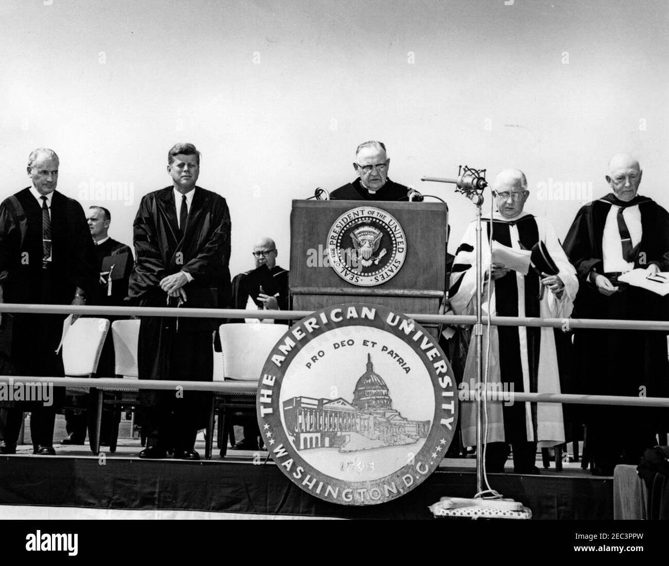 Indirizzo di inizio all'American University, 10:31. Il reverendo Charles R. Smyth, Headmaster della Pennington School di Pennington, New Jersey (al leggio), effettua l'invocazione durante gli esercizi di inizio all'American University. Da sinistra a destra: Il reverendo Warren H. Bright, Jr., Ministro della Chiesa Metodista di Glenwood a Columbus, Ohio; Ralph C. John, Decano del College of Arts and Sciences (in retrocessa); il Presidente John F. Kennedy; non identificato (in retrocessa); il reverendo Smyth; il Dr. Hurst R. Anderson, Presidente dell'Università americana; E John M. Reeves, Presidente del Consiglio di Amministrazione. John M. Reeves Foto Stock