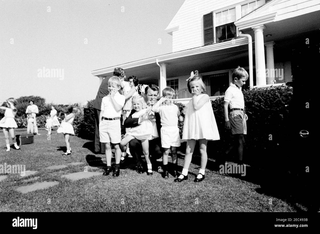 Weekend al porto di Hyannis: Partenza in elicottero e vista aerea del porto di Hyannis, 9:12. Il procuratore generale Robert F. Kennedy si pone con i suoi figli di fronte alla casa dell'ambasciatore Joseph P. Kennedy, Sr., e Rose Fitzgerald Kennedy a Hyannis Port, Massachusetts. In primo piano (L-R): David Kennedy, Kerry Kennedy, Robert F. Kennedy, Michael Kennedy, Courtney Kennedy, e Robert F. Kennedy, Jr. Il presidente John F. Kennedy si trova in background con John F. Kennedy, Jr., e Ann Gargan. Nanny ai bambini Kennedy, Maud Shaw, cammina a sinistra sullo sfondo; altri non sono identificati. Foto Stock