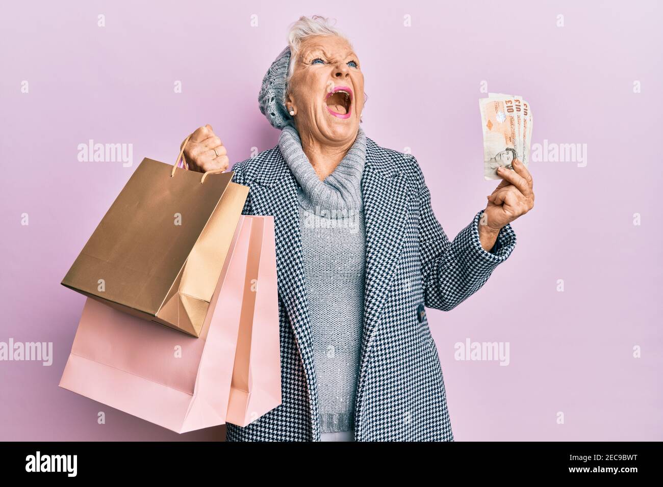 Donna anziana con capelli grigi che tiene borse per la spesa e banconote inglesi in sterline arrabbiate e pazza urlando frustrato e furioso, gridando con rabbia guardando in su. Foto Stock