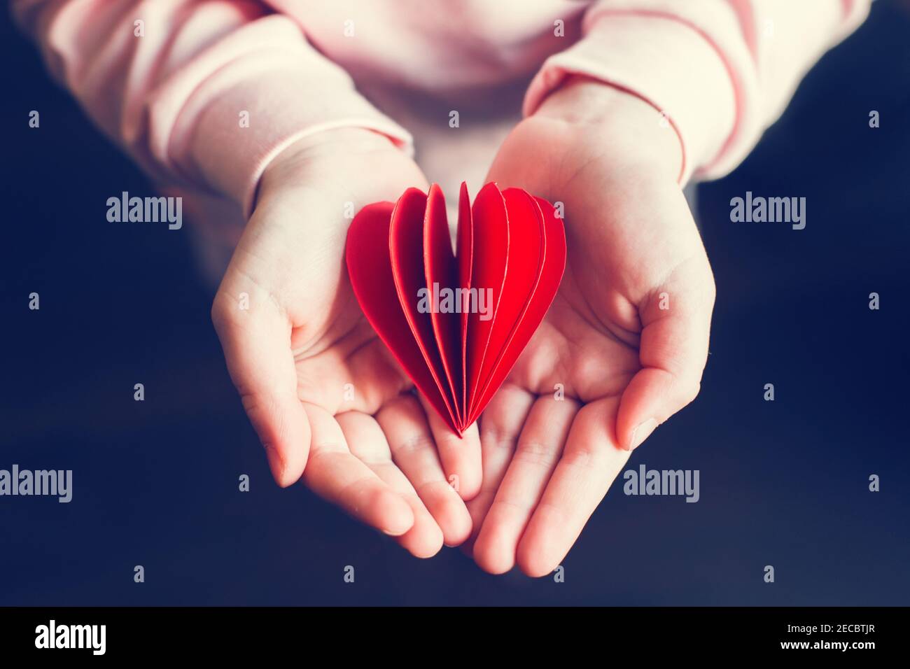 Closeup delle mani del bambino capretto che tengono carta rossa piegata cuore sfondo scuro. Festa di San Valentino. Concetto di supporto, cura e amore. Simbolo del cuore rosso di Foto Stock