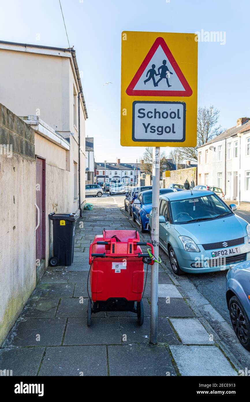 Cardiff, Galles - 12 febbraio 2021: Il trolley di un postista è chiuso ad un cartello di scuola su un marciapiede in una strada Foto Stock