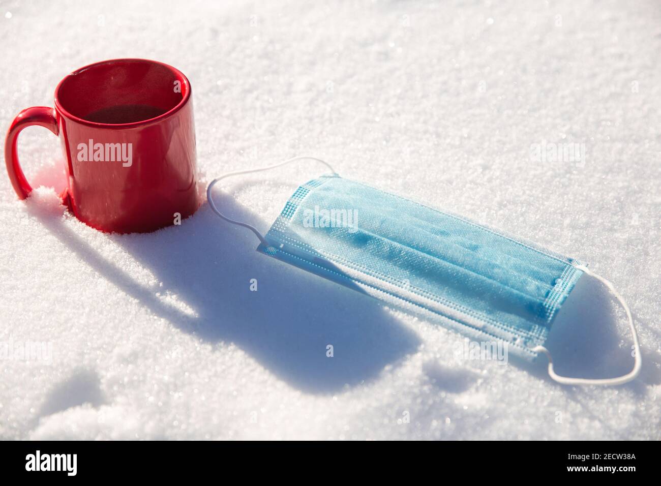 Tazza rossa con caffè caldo, tè o latte di cioccolato e maschera medica blu protettiva per il coronavirus Covid-19 pandemic. Si trova nella neve, inverno, virus, bevanda Foto Stock