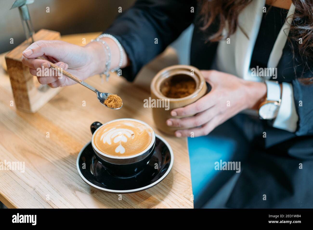 Dall'alto di raccolto anonimo imprenditore femminile in outfit di stile aggiungere un cucchiaio di zucchero di canna in una tazza con cappuccino mentre fare una pausa caffè nel caffè Foto Stock