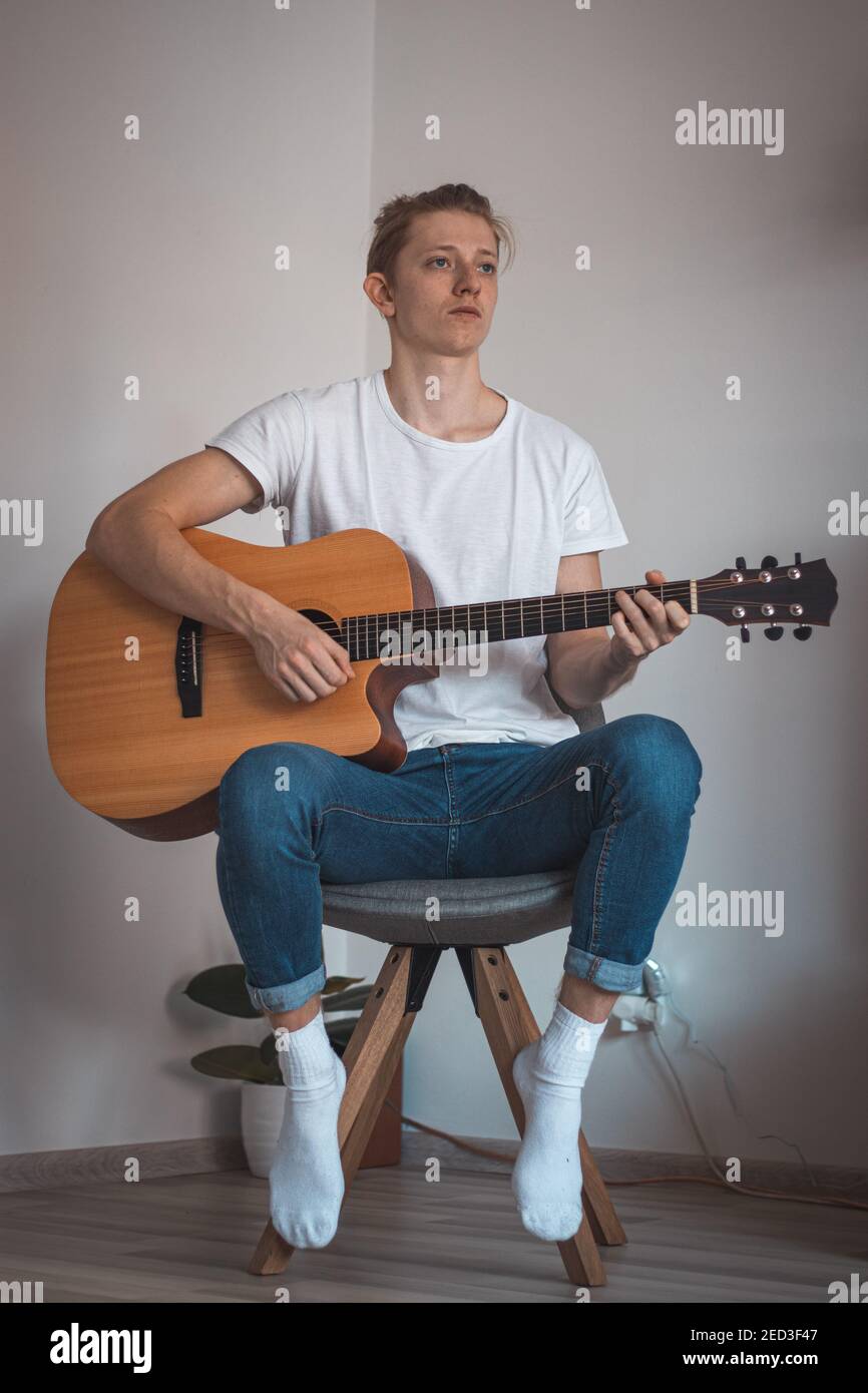 Ritratto candido di ragazzo in una T-shirt bianca e jeans seduti su sgabello basso in angolo della stanza e canta una canzone e suona la sua chitarra. Cerca di trovare inn Foto Stock