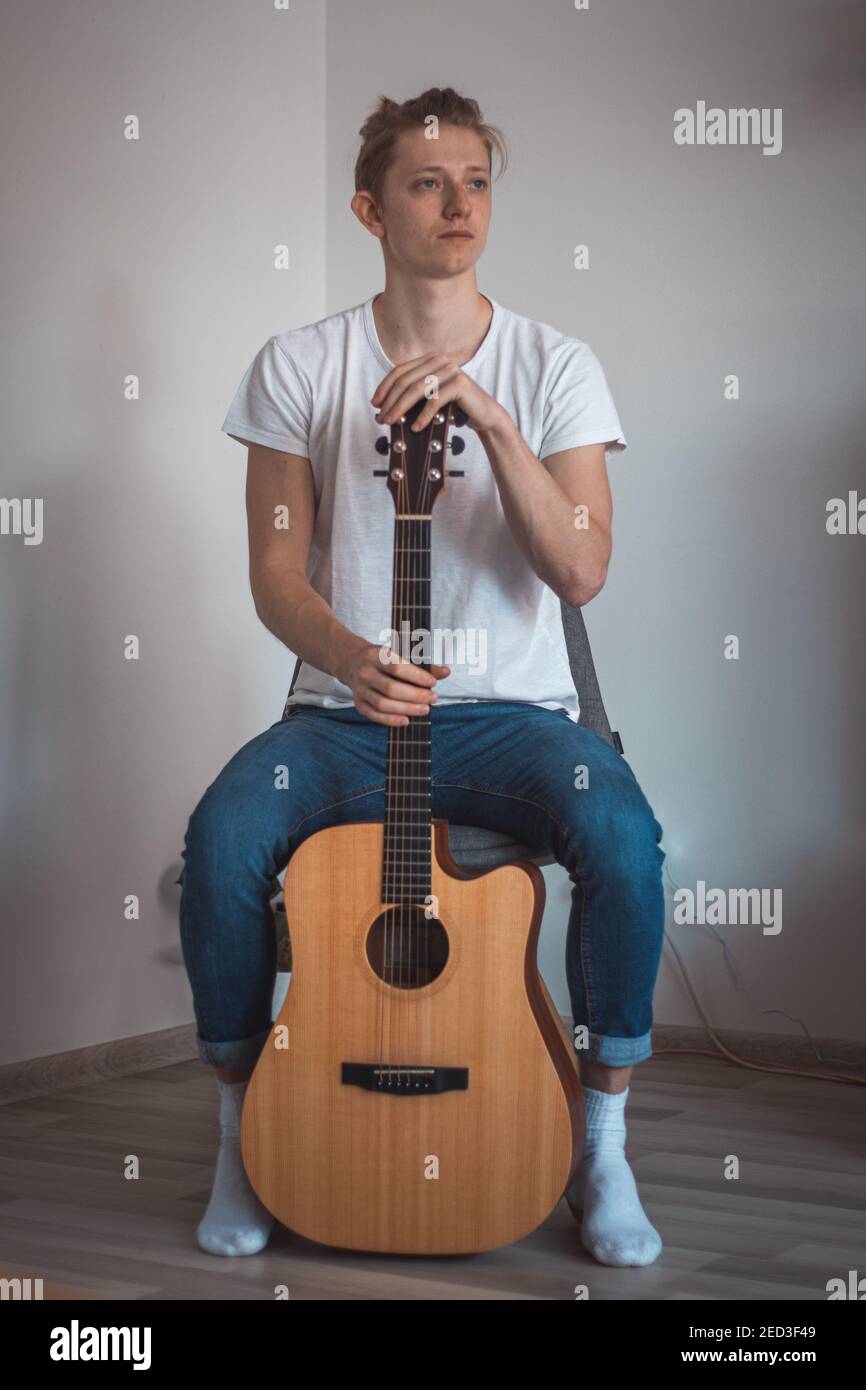 Ritratto candido di un ragazzo in una T-shirt bianca e jeans seduti su uno sgabello basso nell'angolo della stanza e canta una canzone e suona la sua chitarra. Ci prova Foto Stock